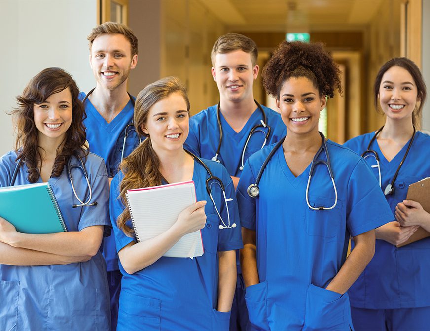 A group of six smiling medical students in blue scrubs stand together in a hallway, holding notebooks and clipboards with stethoscopes around their necks.